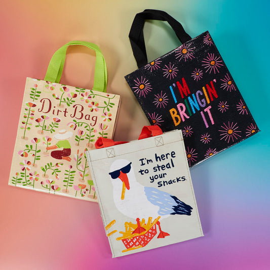 Three colorful reusable tote bags with different designs: one with plants and “Dirt Bag,” one with “I’m Bringin’ It” in bright letters, and one with a seagull and “I’m here to steal your snacks.”.