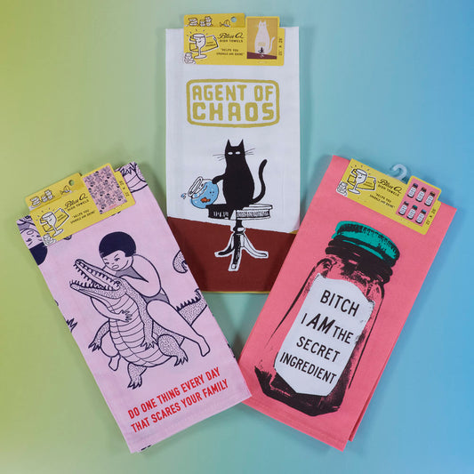 Three colorful dish towels with quirky designs and phrases: a cat labeled Agent of Chaos, a woman wrestling an alligator, and a jar that says Bitch I am the secret ingredient.