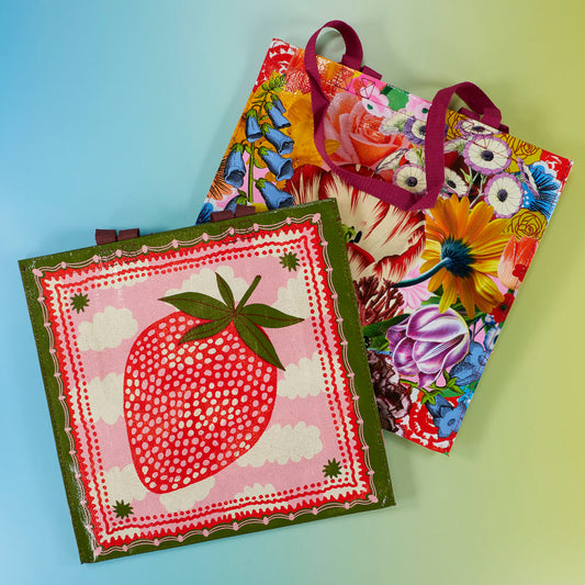 Two colorful reusable tote bags, one with a large red strawberry design on pink and green, and the other with a vibrant floral pattern, placed on a blue-to-yellow gradient background.