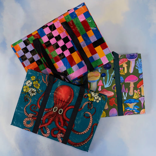 Three colorful reusable tote bags are displayed: one with a red octopus design, one with multicolored checkered squares, and one with various illustrated mushrooms on a blue background.