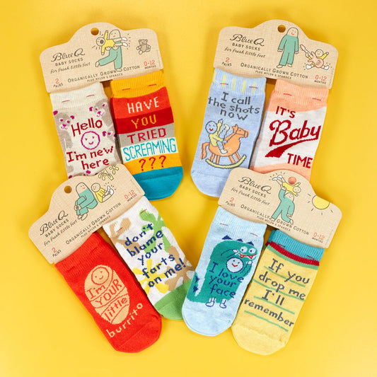 Six pairs of colorful baby socks with humorous phrases, such as “If you can read this, I farted,” displayed on a yellow background. The socks feature cartoonish patterns and text.