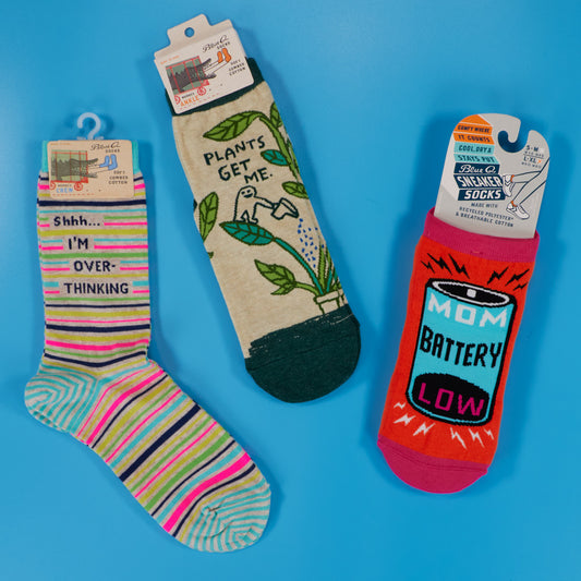 Three pairs of colorful socks on a blue background: one with “Shhh… I’m over-thinking” text, one with a watering can and “Plants get me,” and one with a battery icon labeled “Mom Battery Low.”.