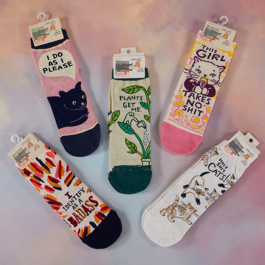 Five pairs of colorful socks with playful cat-themed designs and humorous phrases are arranged in a circle on a pastel background. Each sock has a unique illustration and text.