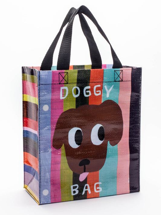 The Blue Q Doggy Handy Tote is a colorful reusable bag with black handles, featuring a cartoon dog's face and "DOGGY BAG" text on a striped background.