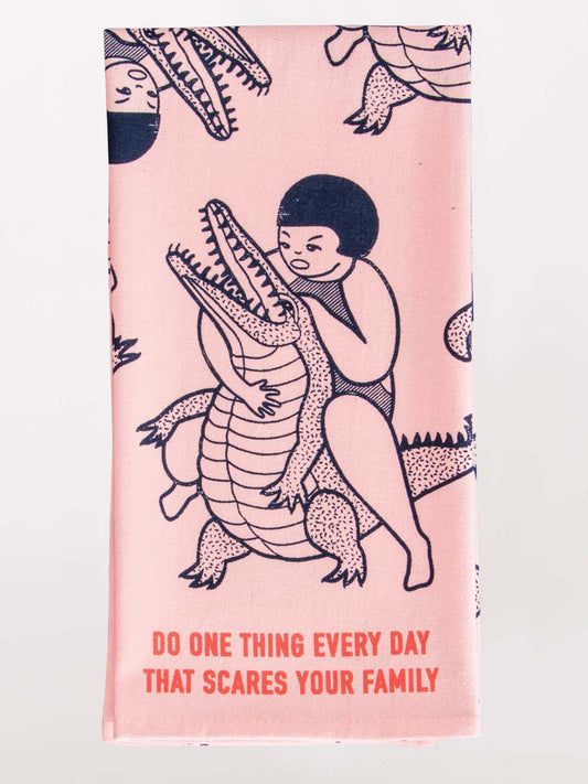 The Blue Q "Do One Thing Every Day That Scares Your Family" Dish Towel is pink, with a person wrestling a crocodile and the motto boldly printed in red at the bottom.