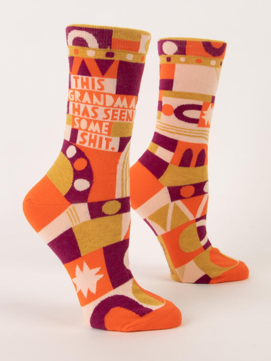 The Blue Q “This Grandma Has Seen Some Shit” W-Crew Socks feature abstract orange, yellow, purple, and cream patterns on soft combed cotton and spandex. One sock displays the quirky phrase; both stand upright against a white background.