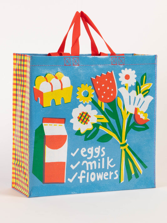 The Blue Q Eggs. Milk. Flowers. Shopper is a reusable bag with red handles, decorated with eggs in a carton, a milk carton, and flowers on blue, plus a checklist: eggs, milk, flowers.