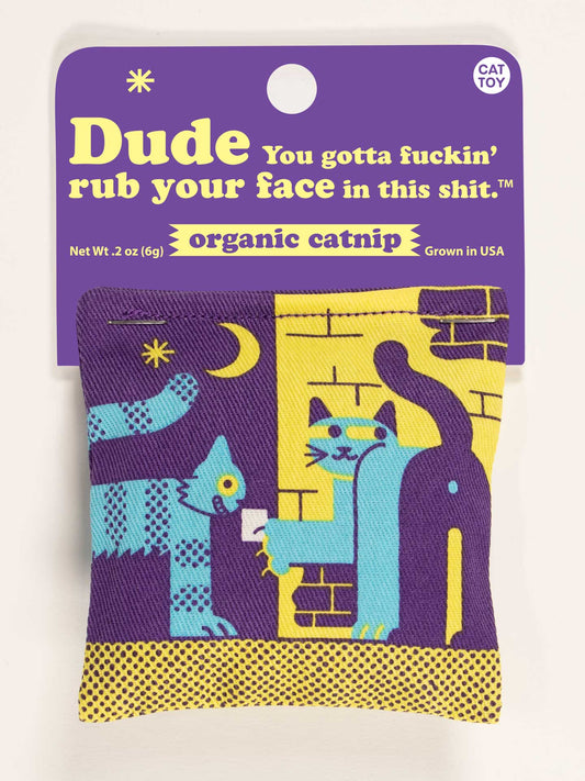 The Blue Q “Dude You Gotta Fuckin' Rub Your Face In This Shit” Catnip Toy is a colorful cotton pouch with a purple and yellow label, filled with organic catnip and decorated with two blue cats in bold geometric patterns.