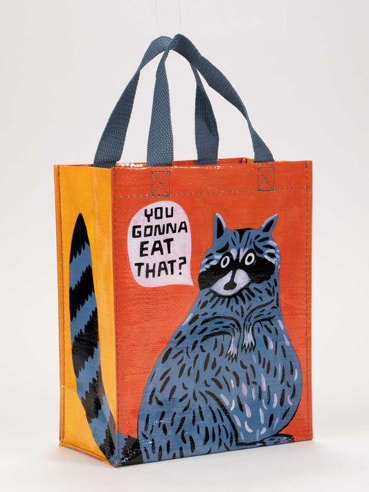 The Blue Q "You Gonna Eat That? Handy Tote" is a reusable orange bag with blue handles, featuring an illustrated raccoon and the fun question: "You gonna eat that?"—perfect for carrying essentials and reducing waste.