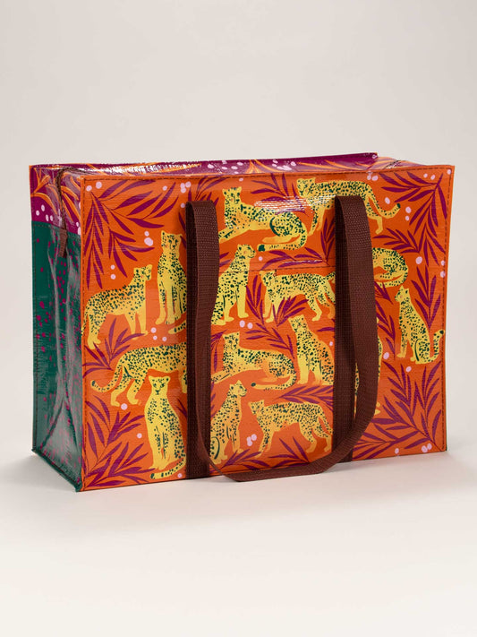 The Blue Q Cheetah Shoulder Tote is a large rectangular reusable bag with brown handles, decorated with yellow cheetahs and purple palm leaves on an orange background.