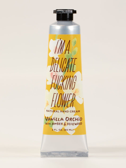 A silver tube of Blue Q's "I'm A Delicate Fucking Flower" Hand Cream – Vanilla Orchid with Amber Rosewood, enriched with shea butter. Decorated with white flowers and a bold yellow label.