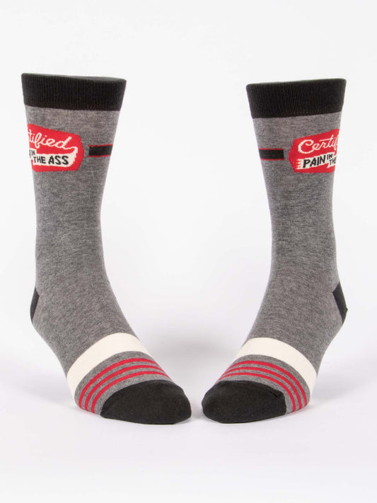 Blue Q’s Certified Pain In The Ass M-Crew Socks are gray combed cotton with black toes and cuffs, red, white, and gray ankle stripes, plus a patch reading “Certified Pain in the Ass.”.