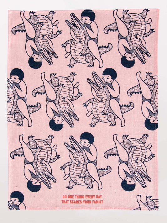 The Blue Q "Do One Thing Every Day That Scares Your Family" dish towel is pink and features a repeating pattern of a child hugging or wrestling a crocodile, with the phrase printed along the bottom.