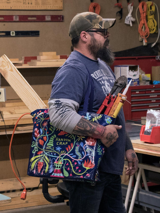 A bearded man in a workshop carries the Blue Q Random Crap Shopper—a colorful tote bag labeled "Random Crap," filled with tools and a wooden plank—surrounded by workshop equipment, ideal for groceries or gear.