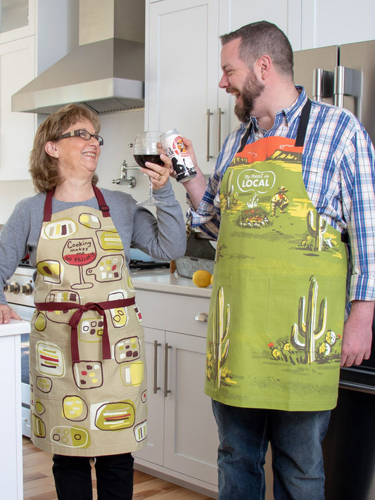 A happy couple wearing Blue Q "My Meat Is LOCAL" aprons with extra-long waist ties stand in a modern kitchen, holding drinks—a wine glass and a canned beverage—cheering together and looking relaxed.