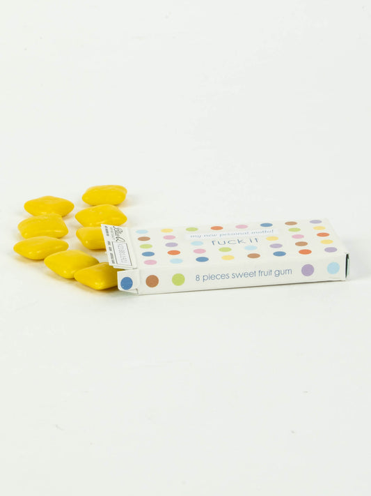 A rectangular box labeled "Fuck It Gum" by Blue Q, showing 8 sweet fruit flavor pieces with colorful dots, partially open and yellow oval gum spilling out like classic French candy onto a white surface.