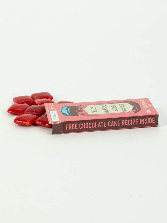 A pink rectangular box labeled "Bitch I Am The Secret Ingredient Gum" by Blue Q lies on its side, partially open, with several red, pillow-shaped cinnamon-flavored gum pieces spilling onto a white surface.