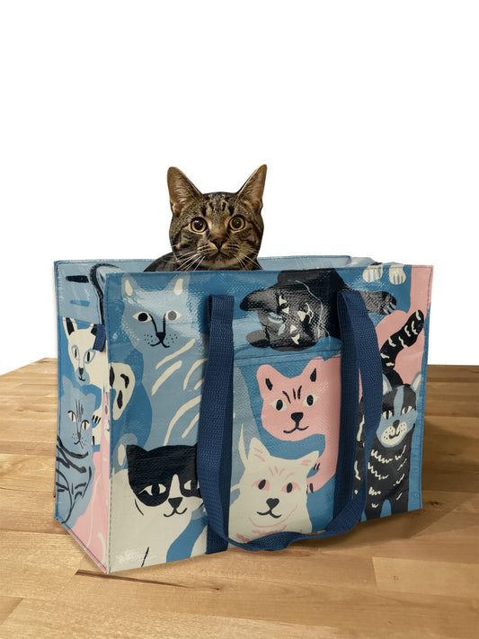 A brown tabby cat sits inside the Blue Q Happy Cats Shoulder Tote, a comfy bag with colorful cartoon cat faces, on a wooden surface against a white background.