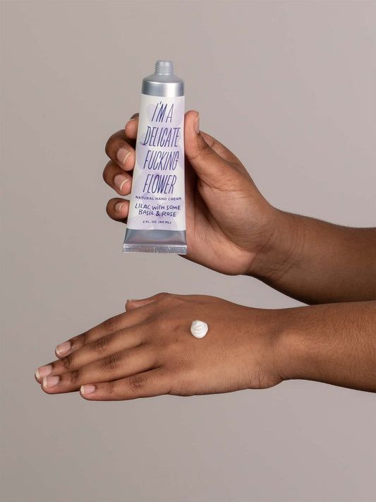 A person applies a small amount of Blue Q's I'm a Delicate Fucking Flower Natural Hand Cream - Lilac with Some Basil & Rose onto the back of their hand, with the tube visible against a neutral background.