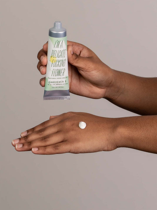 A person applies Blue Q's I'm a Delicate Fucking Flower Natural Hand Cream - Gardenia & a Bit of Sandalwood onto the back of their hand, with a small dollop of cream visible on their skin.