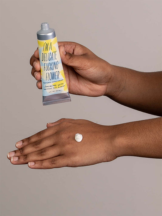 A person squeezes Blue Q’s I’m a Delicate Fucking Flower Natural Hand Cream—Jasmine with Orange Peel & Pear—from a tube onto the back of their hand against a plain background.