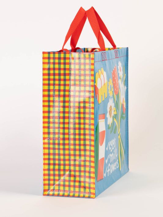 The Blue Q "Eggs. Milk. Flowers. Shopper" is a reusable shopping bag with red handles, a colorful plaid side, and playful illustrations of eggs, milk, flowers, and a grocery list on the blue front panel.