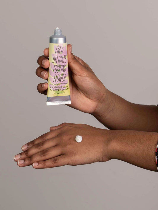 A person applies Blue Q's I'm a Delicate Fucking Flower Natural Hand Cream—lavender with a little lemon, infused with almond oil and shea butter—from the tube to the back of their hand.