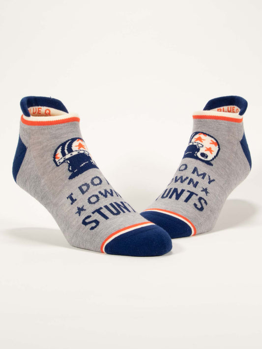 Blue Q's I Do My Own Stunts Sneaker Socks are gray ankle socks with blue toes and heels, orange accents, and a helmet-and-stars design. They feature shock-absorbing cushion and display the text "I DO MY OWN STUNTS.