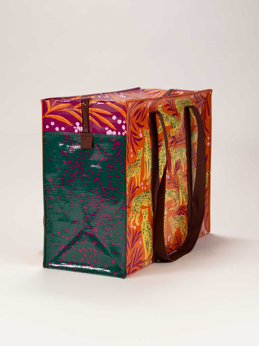 The Blue Q Cheetah Shoulder Tote is a colorful reusable bag with brown handles, featuring a green panel with pink dots on one side and orange panels adorned with bold cheetah and leaf prints on the others.