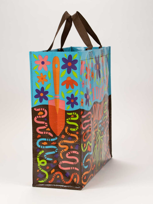 The Blue Q I Dig Dirt Shopper is a colorful reusable bag with brown handles, featuring a garden trowel, flowers, roots, and cartoon worms on bright blue, green, and brown soil-inspired backgrounds.