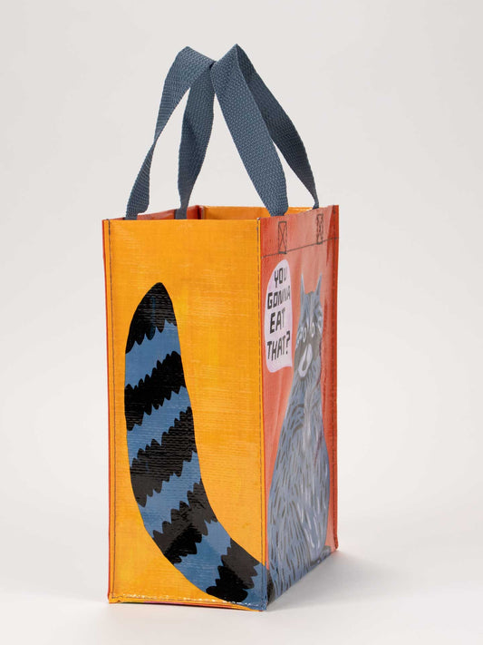 The Blue Q "You Gonna Eat That? Handy Tote" features blue handles, orange sides, and a painted raccoon asking "You gonna eat that?" in a speech bubble, with its striped blue and black tail on one side.