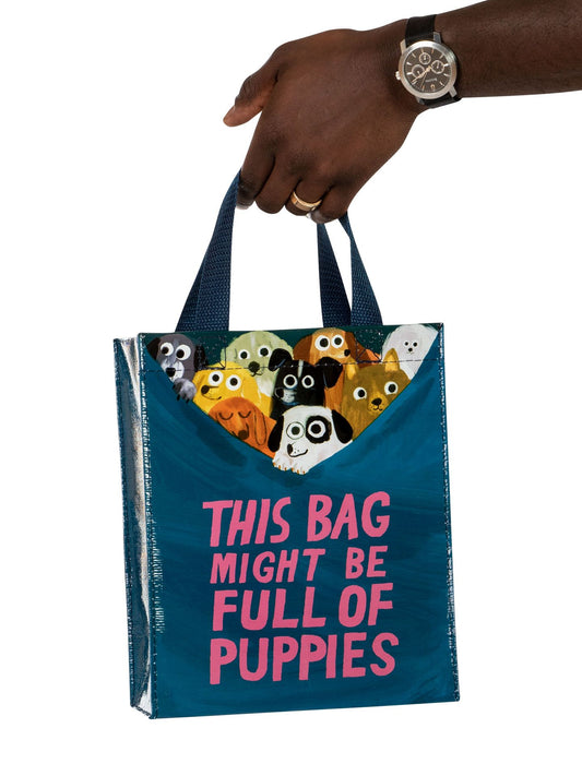 A hand holds Blue Q’s "This Bag Might Be Full Of Puppies Handy Tote," a blue tote with cartoon puppy faces and bold pink text—perfect for dog lovers who’d rather carry pups than $50,000.