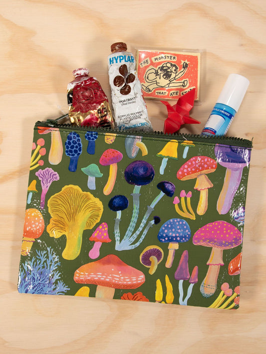 The Blue Q Mushrooms Zipper Pouch, featuring a colorful mushroom design, is shown unzipped on a wooden surface with paint tubes, a small book, and other items neatly stored inside.