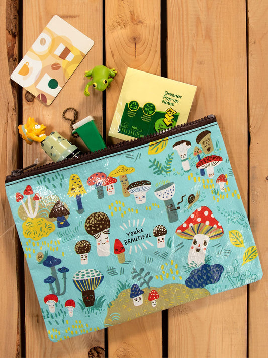 The Blue Q Cute Lil Mushrooms Zipper Pouch, featuring whimsical mushrooms and animals with a "You're Beautiful" label, sits on a wooden surface near a frog keychain, lighter, notepad, novelty pencil toppers, and a patterned card.