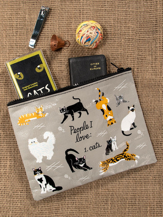 The Blue Q "People I Love: Cats Zipper Pouch," featuring cartoon cats and the phrase “People I love: 1. cats,” sits on burlap surrounded by nail clippers, a jingle bell, a rubber band ball, a passport case, and a cat-themed notebook.