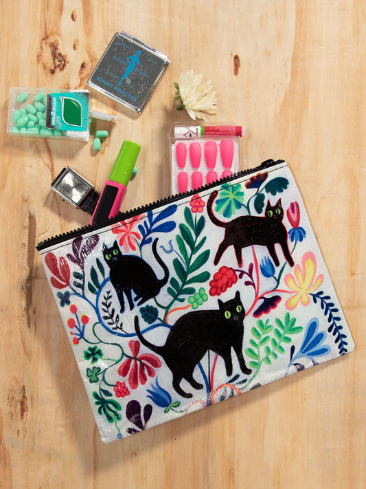 The Blue Q Here Kitty Zipper Pouch, featuring a black cat and floral design, rests on a wooden surface alongside cosmetics, teal pills, and fake pink nails.