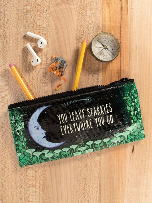The Blue Q "You Leave Sparkles Everywhere You Go" Pencil Case with a moon design sits on a wooden surface among glitter gel pens, a sharpener with shavings, a compass, and wireless earbuds.