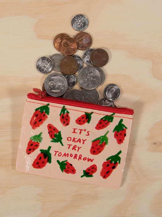 The Blue Q It's Okay Try Tomorrow Coin Purse, decorated with strawberries and an uplifting message, is open on a light wood surface with coins spilling out—ideal for saving up for your next frozen yogurt treat.