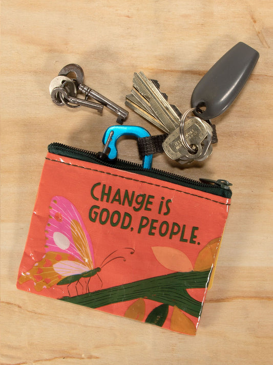 A set of keys and a gray key fob are attached to the Blue Q Change Is Good, People. Coin Purse with a butterfly design, resting beside nachos on a light wooden surface.