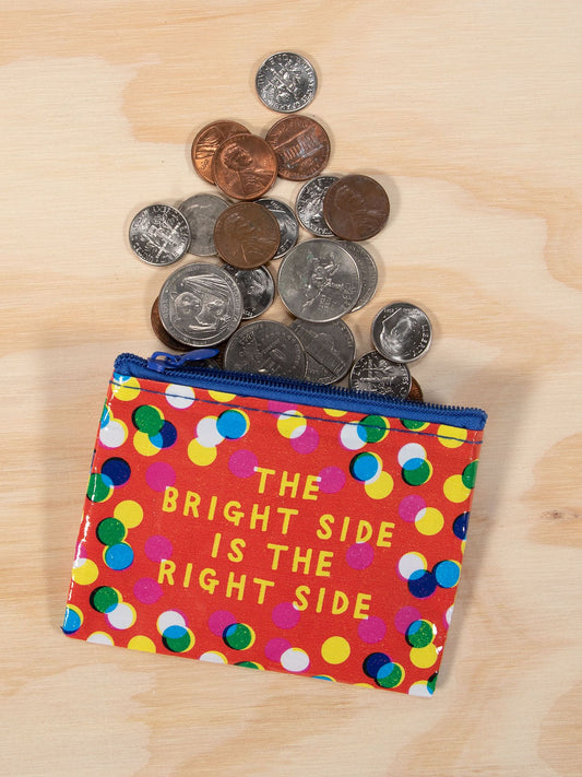 The Blue Q "The Bright Side Is The Right Side" Coin Purse features a blue zipper and polka dot design, adding cheerful vibes as assorted coins spill onto the light wood surface.
