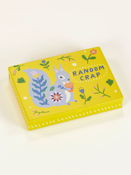 The Blue Q Random Crap Pocket Box is a yellow storage box with colorful flowers and a cartoon squirrel holding an acorn. The lid reads "RANDOM CRAP" in white, making it a fun way to organize your small items.