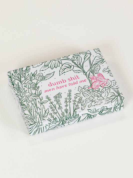 The Blue Q "Dumb Shit Men Have Told Me Pocket Box" features green floral line art, a pink butterfly, and bold pink text on a rectangular lid—ideal for collecting memorable quotes. The background is light and neutral.
