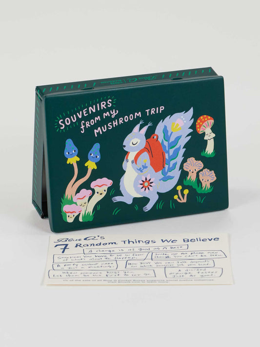The Blue Q Souvenirs From My Mushroom Trip Pocket Box features a whimsical squirrel with a red backpack among colorful mushrooms, plus sparkle pebbles and a handwritten note card on the illustrated box.