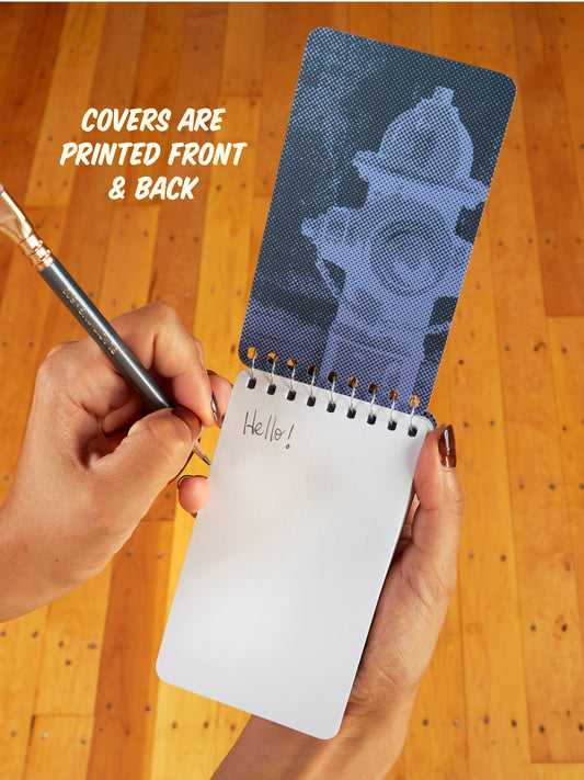 Using the Blue Q "I Love To Go With The Flow. My Flow, Not Yours" 3x5 spiral notebook with a blue cover and fire hydrant, someone writes "Hello!" as “COVERS ARE PRINTED FRONT & BACK” glows above on a wooden floor.