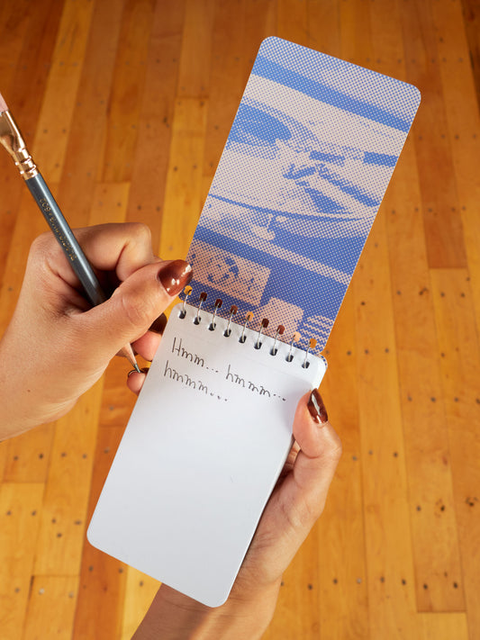 A person writes "Hmm... hmmm... hmmm..." with a pencil in the Blue Q "I've Said It Before And I'll Say It Again And Wait I Forgot" 3x5 Spiral Notebook. Their nails are painted brown, and a wooden floor is in the background.