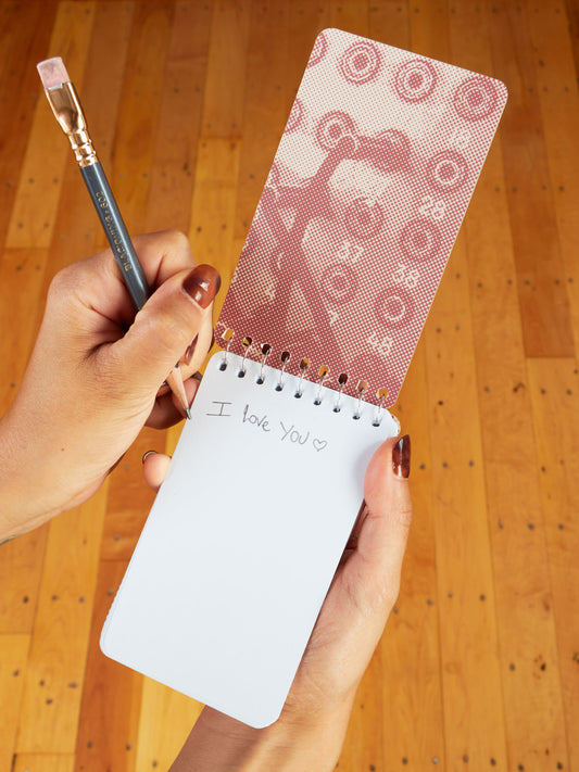 A person holds the Blue Q "Just Between You And Me I'm Gonna Tell Everyone" 3x5 Spiral Notebook and a paintbrush. On its open page, "I love you" with a heart is written in pencil against a wooden floor background.