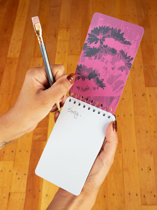 Someone writes "Sorry." in a Blue Q "Sorry For Being Rude. I Have This Thing Where I'm Rude" 3x5 spiral notebook featuring a pink and black stylized face cover, using a pencil with a paintbrush instead of an eraser.