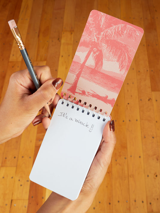 A person with brown nail polish writes "It’s a miracle!!" in a Blue Q "Took A Taste Of My Own Medicine And Would You Look At That I'm Cured" 3x5 Spiral Notebook with a pink palm tree cover, sitting on a wooden floor.