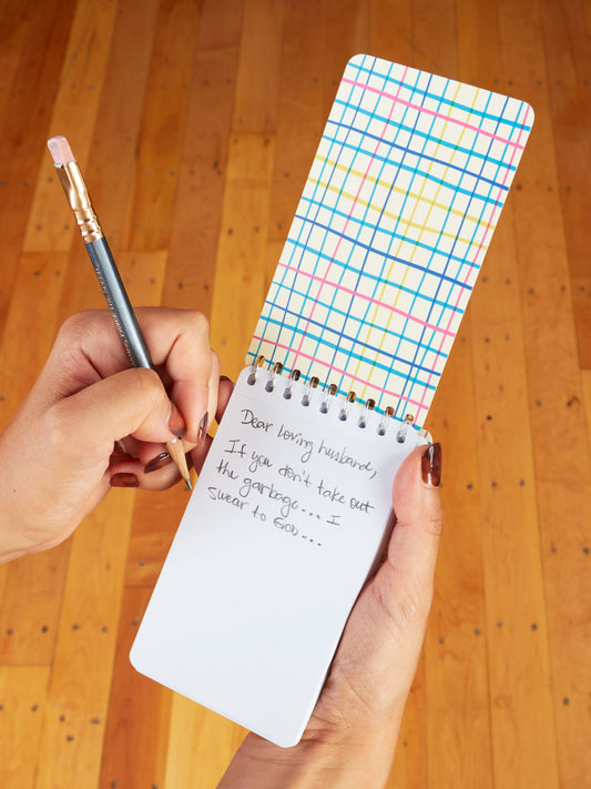 Using a Blue Q "Sweet Little Reminders For My Stupid Husband" 3x5 spiral notebook, someone jots a funny love note: "Dear loving husband, if you don’t take out the garbage… I swear to God..."—a quirky romantic gift.