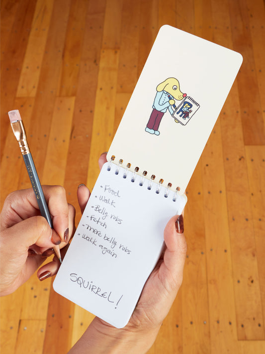 A hand writes in the Blue Q "Diary Of A dog. Licked Some Faces, Sniffed A Bush, Barked At A Vacuum" 3x5 spiral notebook featuring a cartoon dog with a clipboard and a to-do list: Food, walk, belly rubs, SQUIRREL! on a wooden floor background.